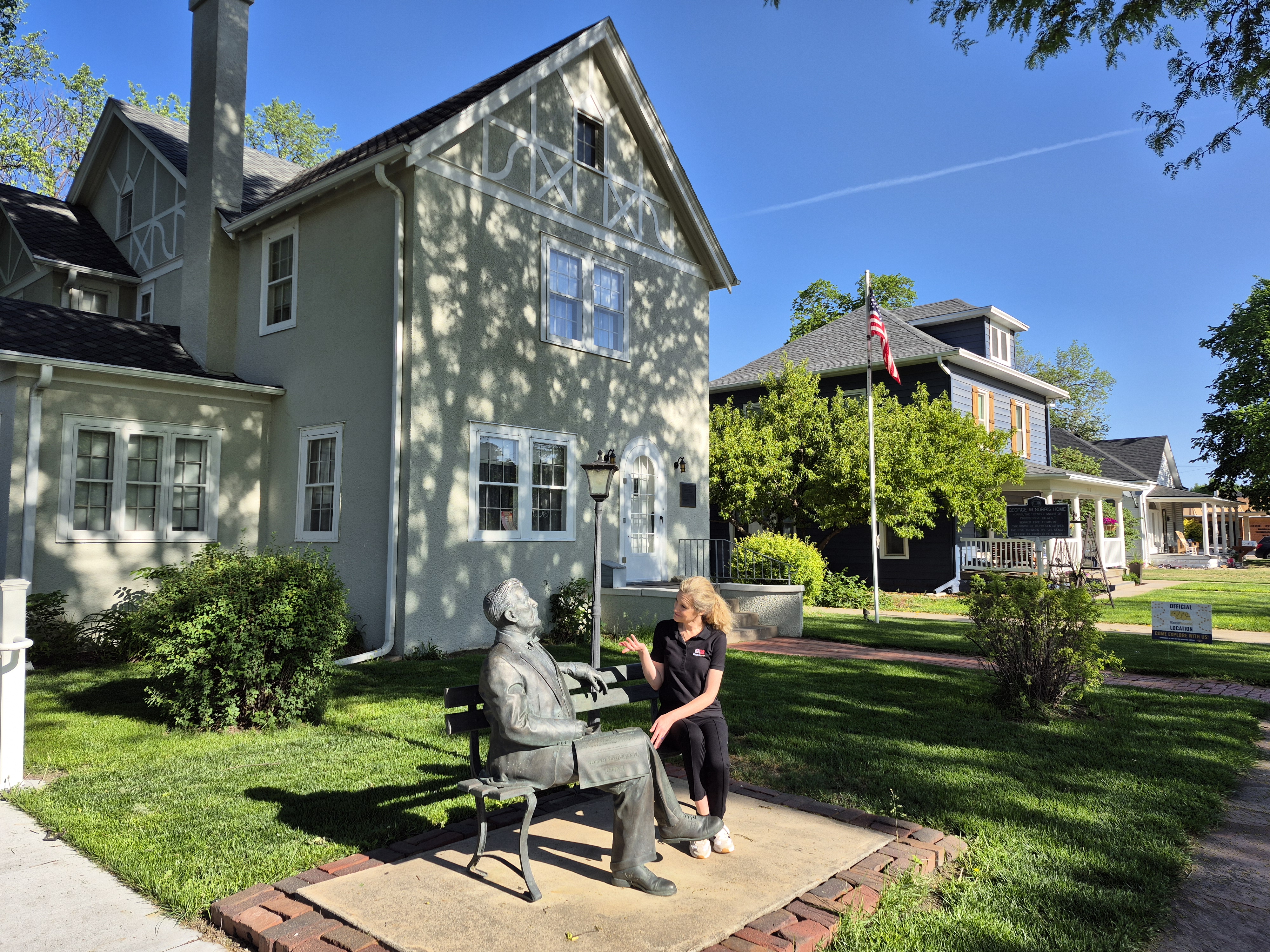 Woman sitting next to a bronze statue on a bench outside historic houses under a sunny blue sky.