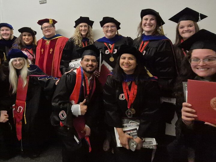 Graduates in caps and gowns holding diplomas and posing in a hallway.